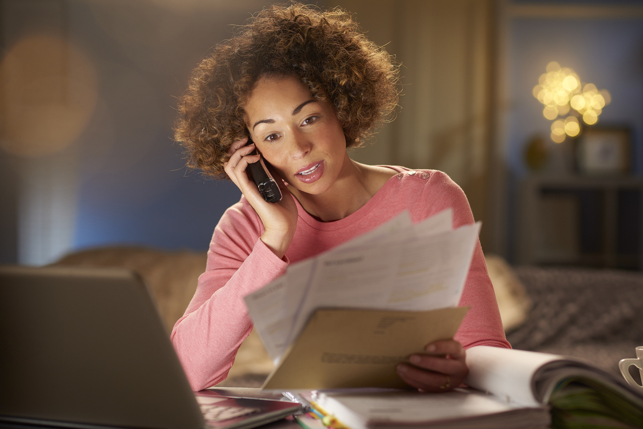 Young woman sits at home, going through her paperwork and bills. She is working at her laptop and appears concerned as she speaks to someone on the phone about her bills.