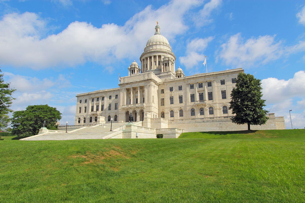 Front view of the Rhode Island state capitol building and lawn on Capitol Hill in Providence with bright blue sky and white clouds in the background. The building is covered with white Georgia marble and was constructed from 1895 to 1904.